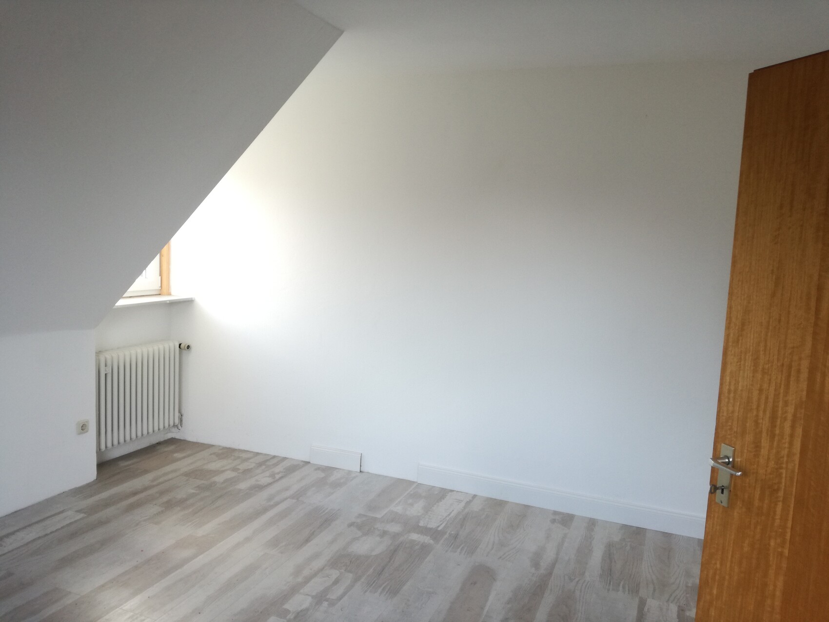 Kleines, leeres Dachgeschosszimmer mit schräger Decke, Fenster, Heizkörper und Holzboden.