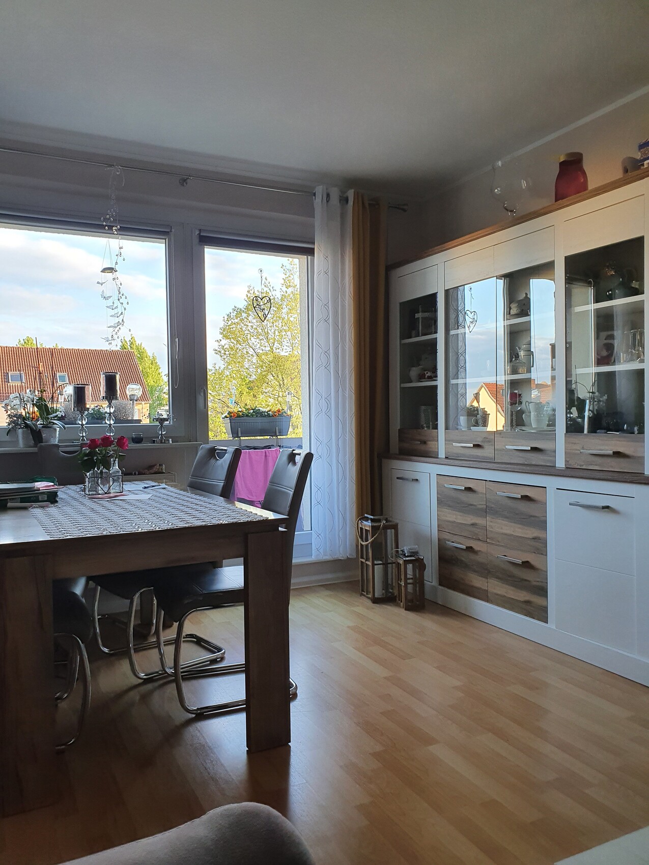 Heller Essbereich mit Holztisch, Glasvitrine und Fensterfront; draußen Häuser und Bäume sichtbar.