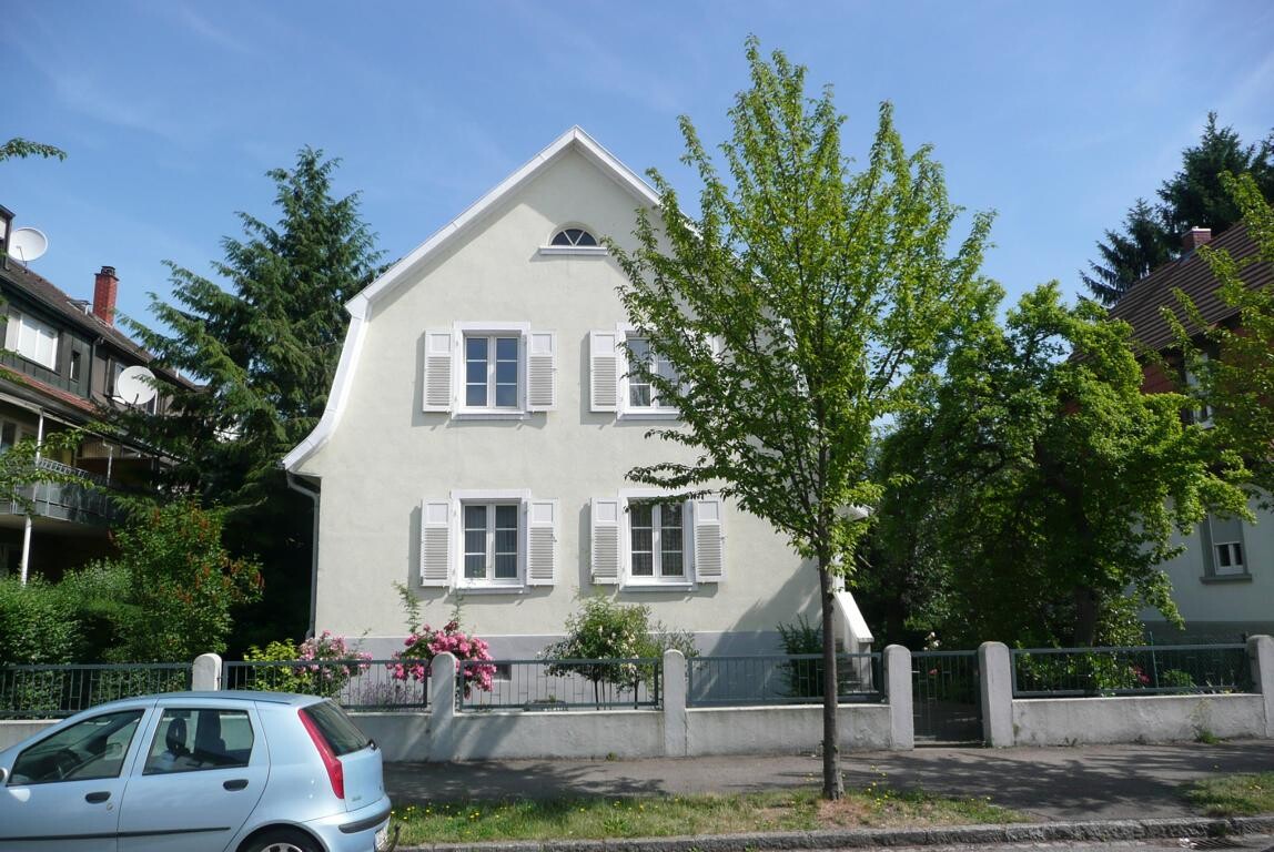 Außenansicht eines hellen Hauses mit weißen Fensterläden, Baum und Zaun