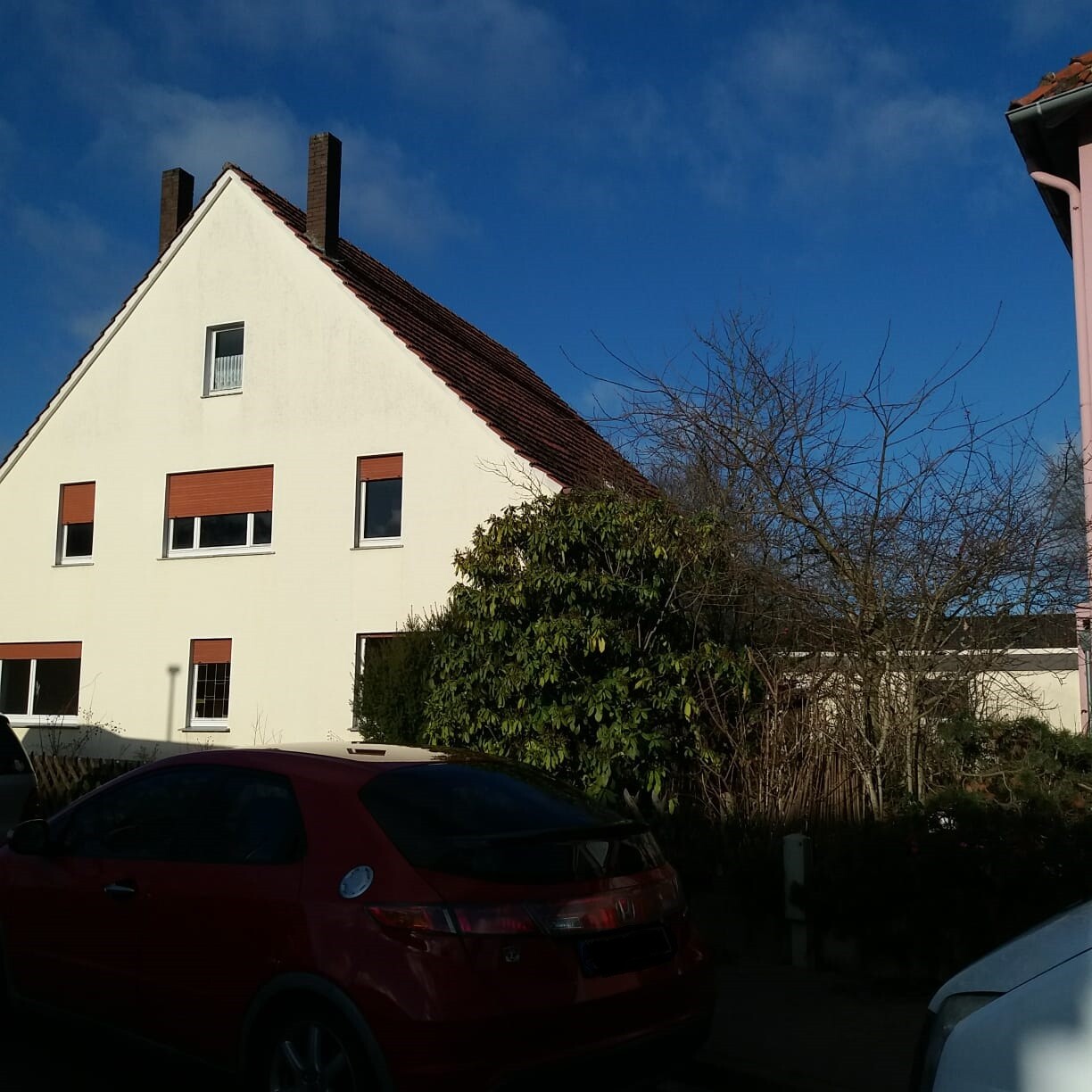 Weißes Haus mit rotem Ziegeldach, Fenstern mit Rollläden, Busch und rotem Auto davor.