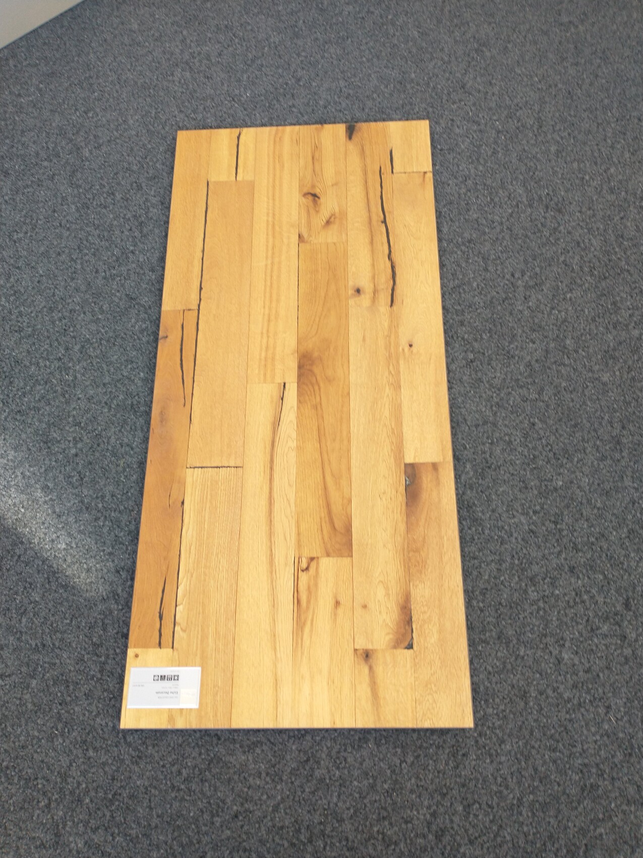 Holzplatten-Bodenprobe aus hellen Dielen auf grauem Teppich, Etikett unten links.