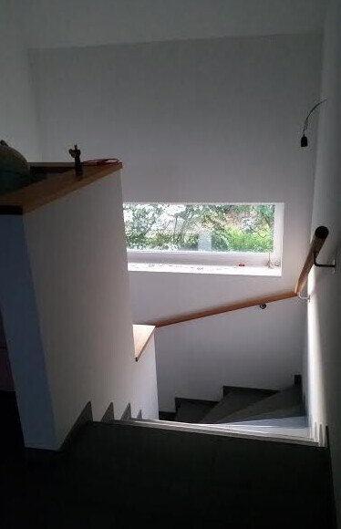 Innenaufgang einer Treppe mit Holzhandlauf, weißer Wand, kleinem Fenster mit Blick nach draußen.