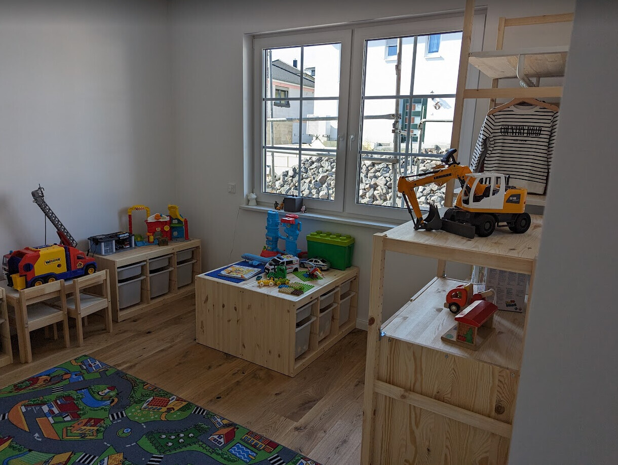 Spielzimmer mit großem Fenster, Holzregalen, Spielzeugfahrzeugen und bunter Spielmatte