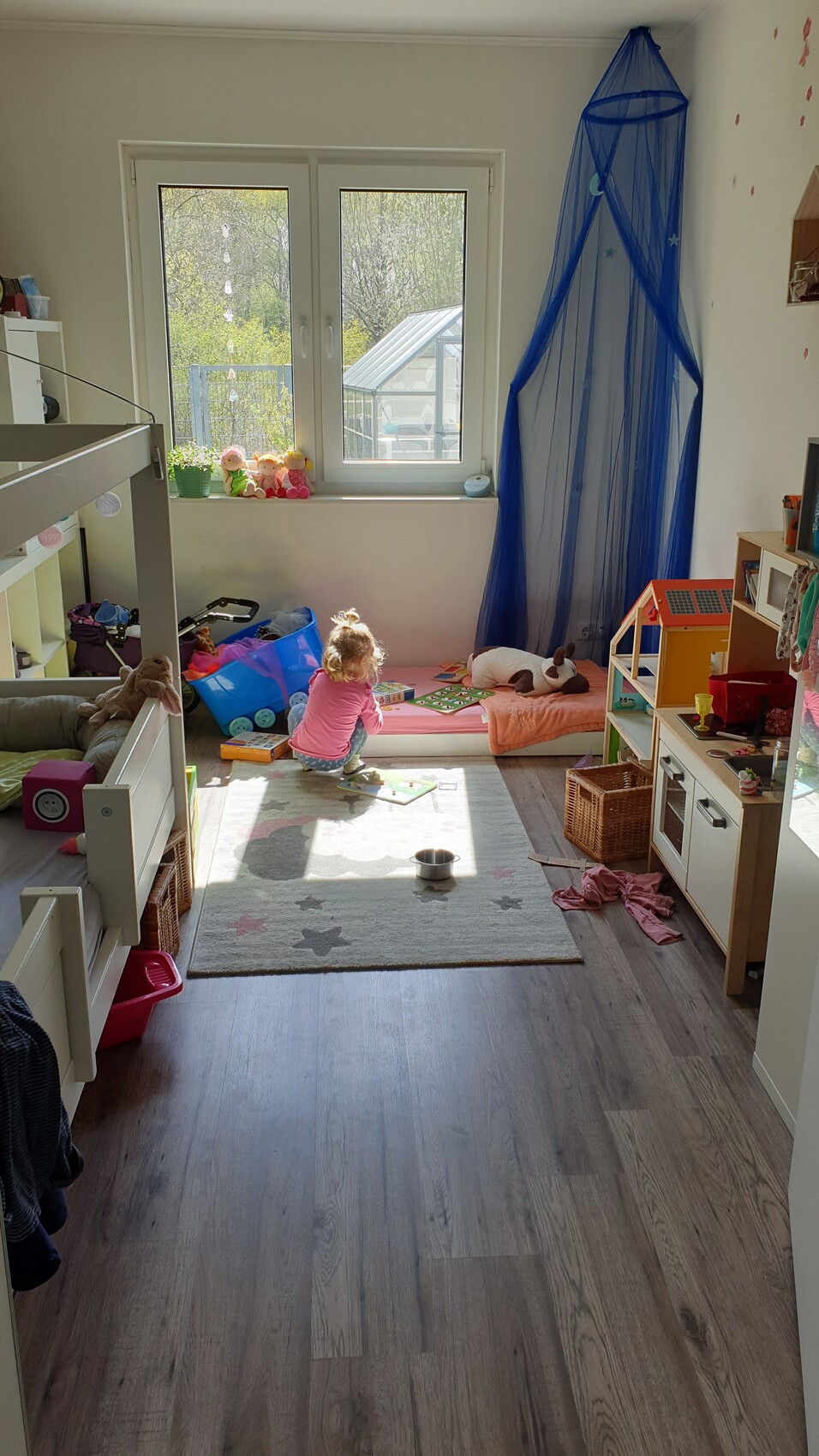 Kleines Kind spielt auf Spielmatte im hellen Kinderzimmer; Bett, Spielzeug und blauer Netzvorhang.