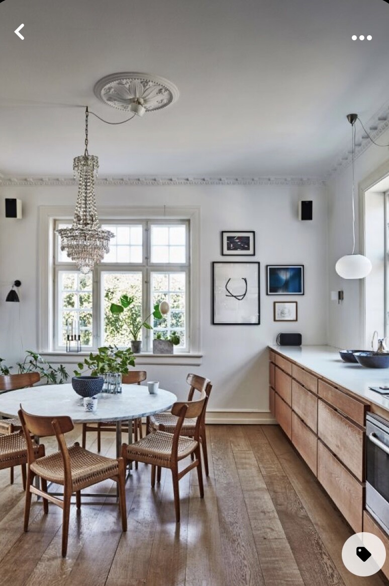 Helles Esszimmer: runder Marmortisch, Holzmöbel, Kronleuchter, Fensterpflanzen, Küchenzeile.