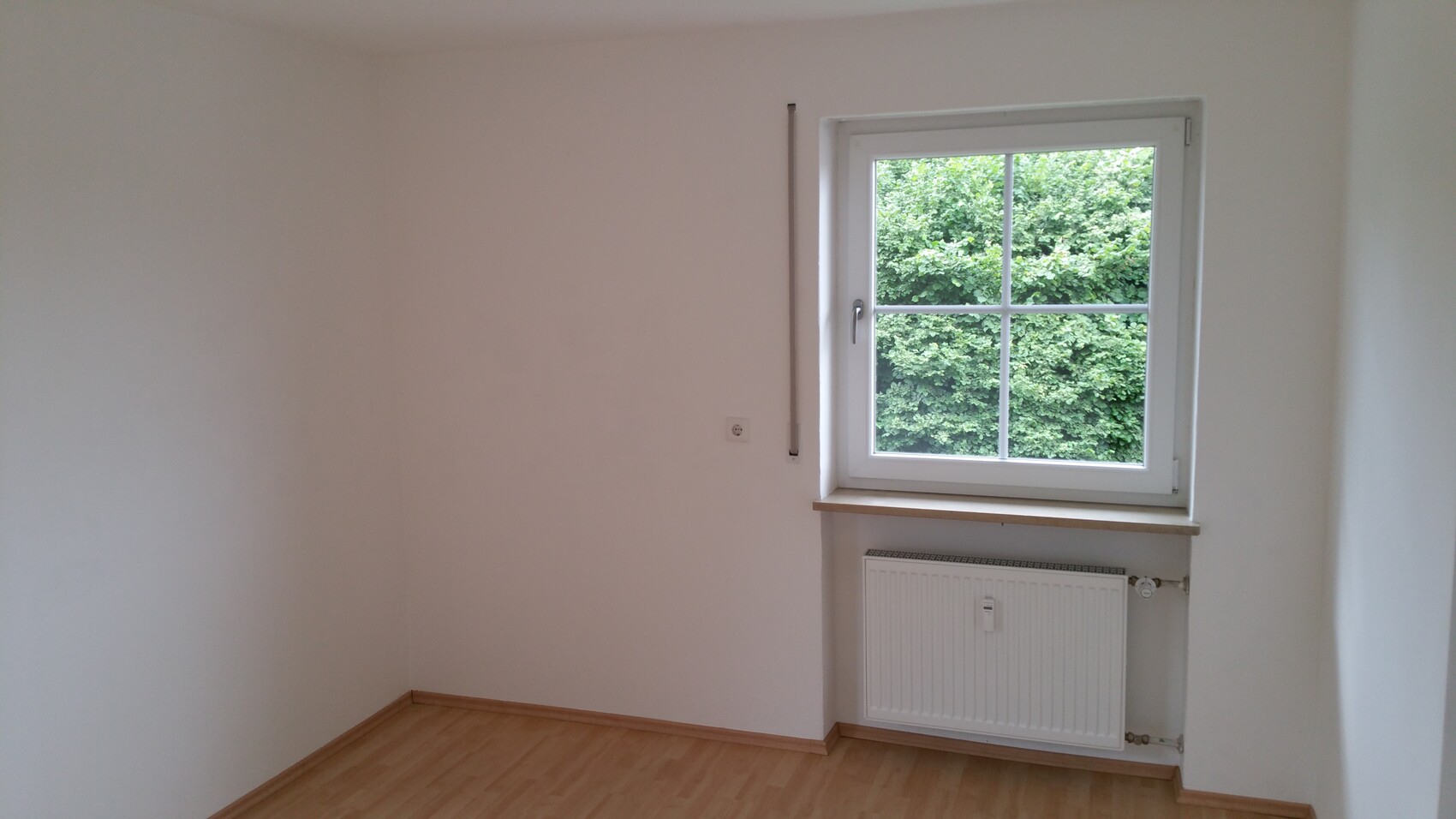 Kleines, leeres Zimmer mit Fenster, Holzboden, Heizkörper unter dem Fenster und Gartenblick.
