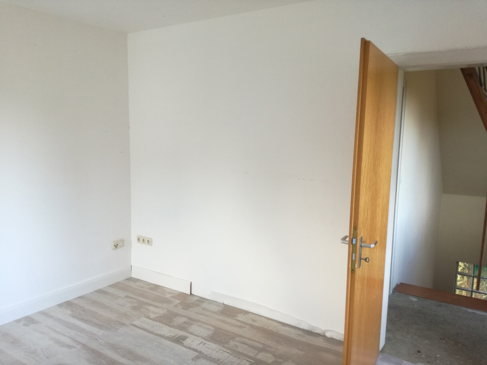 Leerer Raum mit weißen Wänden, offener Holztür, Laminatboden und Steckdosen.