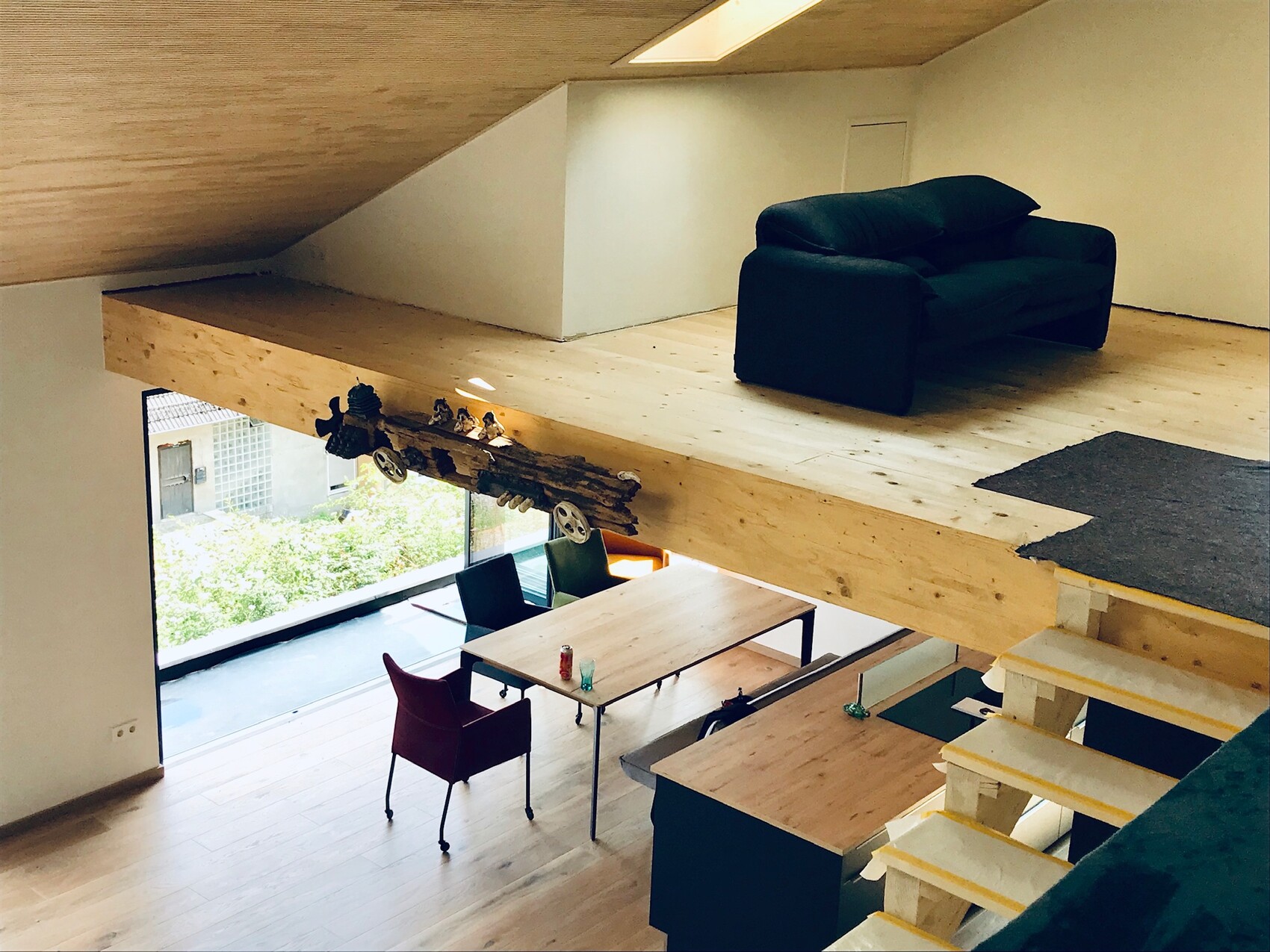 Loft mit Holzboden und Balken, obere Lounge mit schwarzem Sofa, Esstisch unten, großes Fenster.