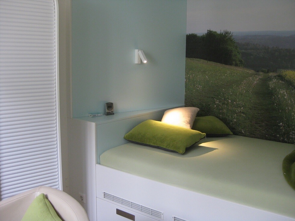 Schlafzimmer mit hellgrüner Bettwäsche und zwei Kissen; Wandbild einer Wiesenlandschaft mit Pfad.