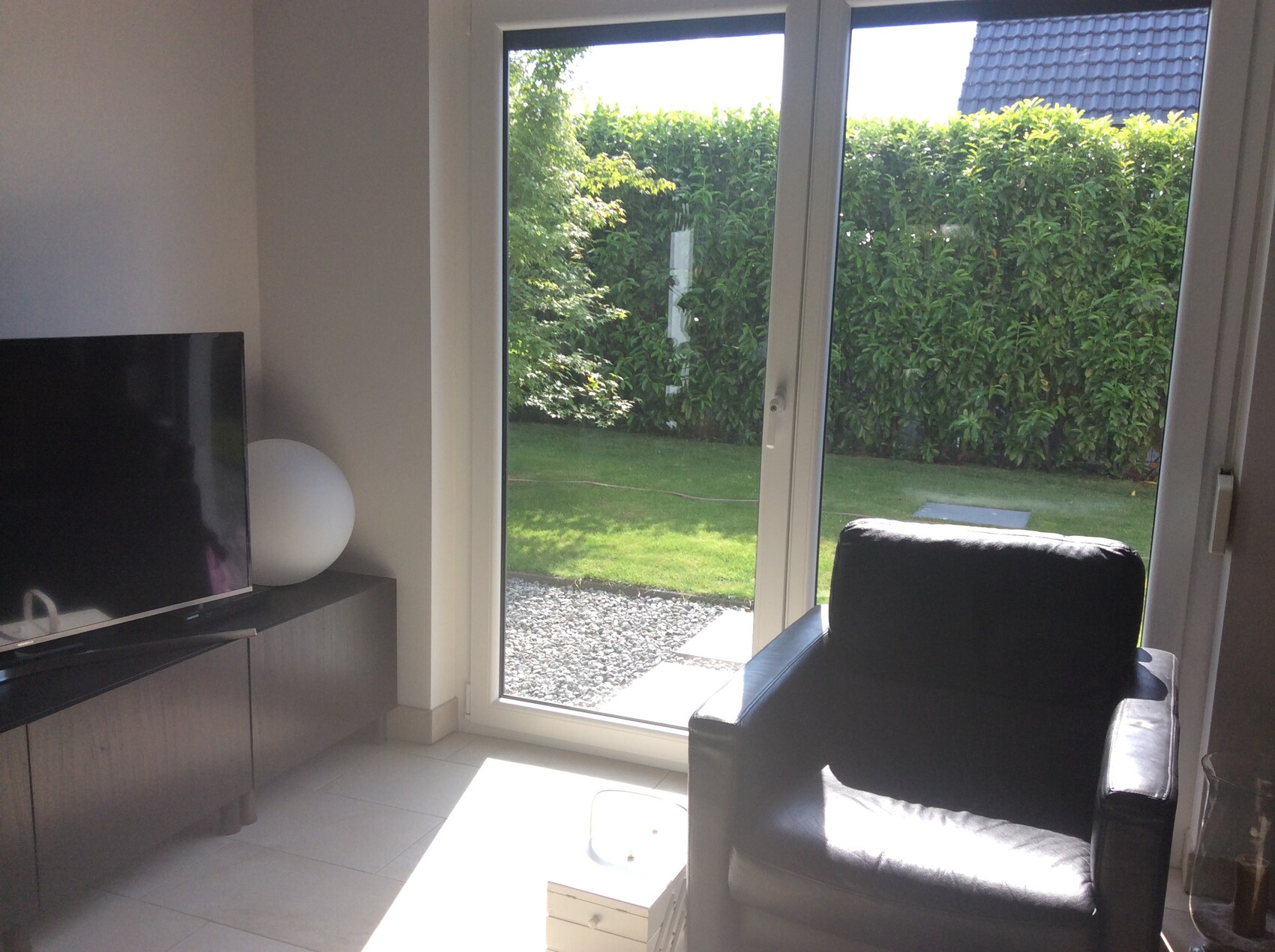 Wohnzimmer mit schwarzem Ledersessel vor Glas-Schiebetür zum grünen Garten; TV auf Sideboard.
