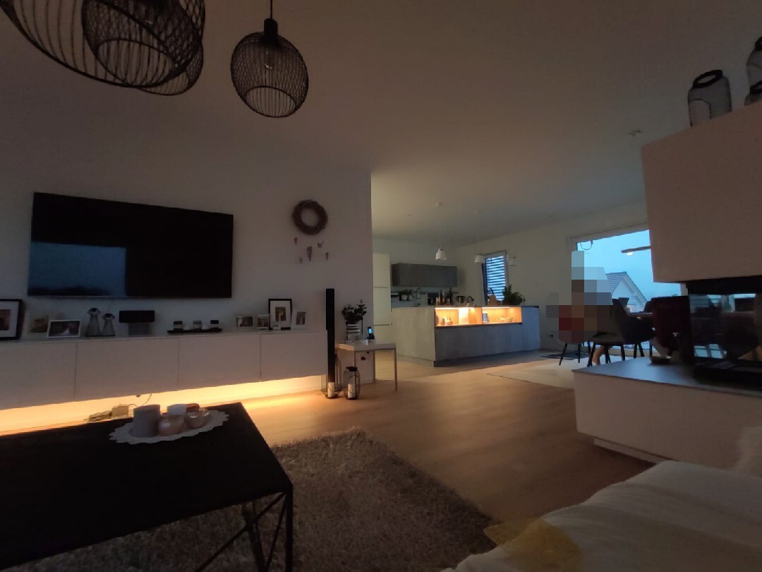 Moderner offener Wohnraum mit TV-Wand, weiße Möbel, offene Küche im Hintergrund, Tisch mit Kerzen.