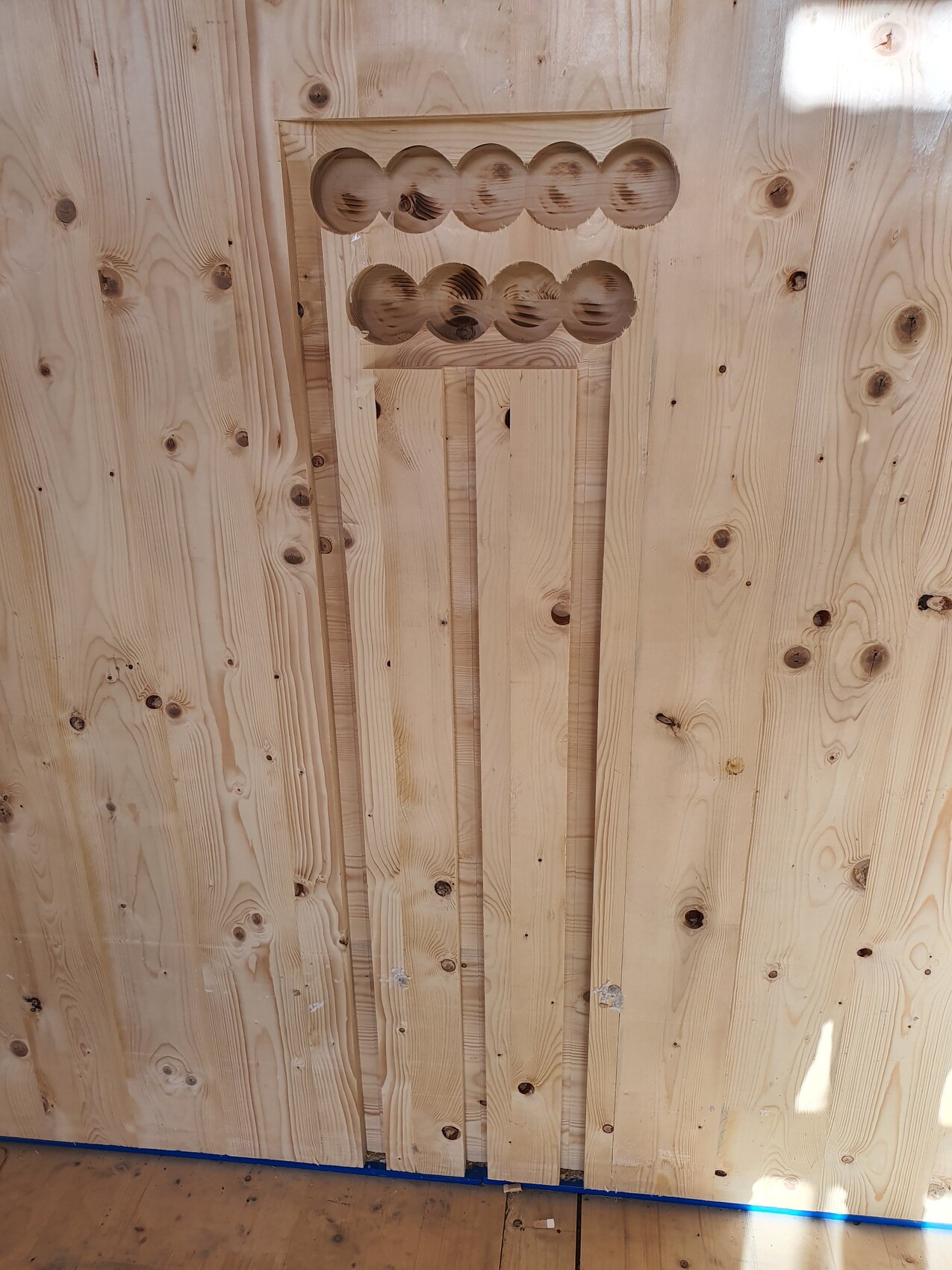 Holzwand aus Brettern; zentrales Relief mit zwei Reihen runder Vertiefungen.