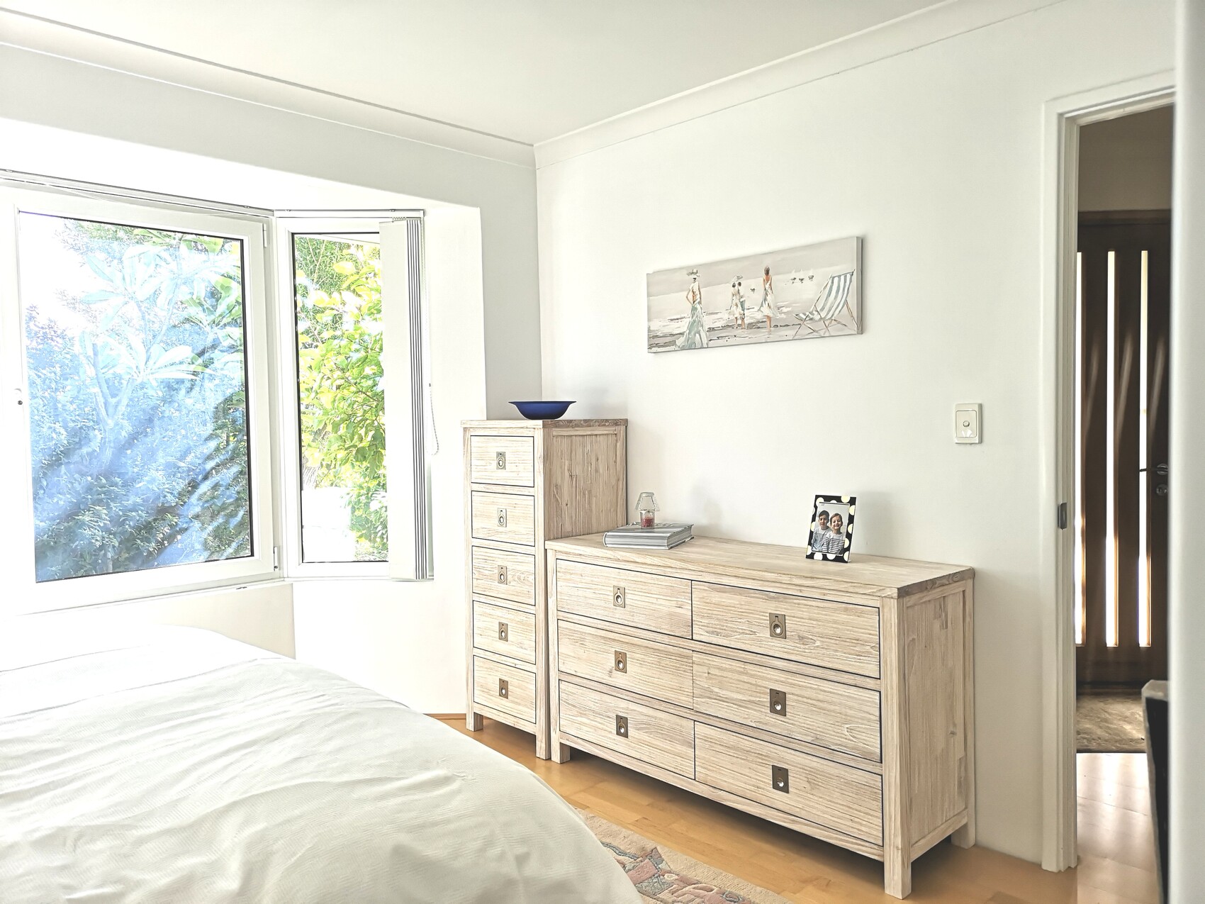 Helles Schlafzimmer mit offenem Fenster, Bett links, helle Holzmöbel und Strandbild an der Wand.