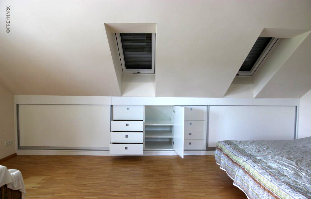 Schlafzimmer mit weißer Schrankwand unter Dachschrägen und Dachfenstern; Bett rechts.