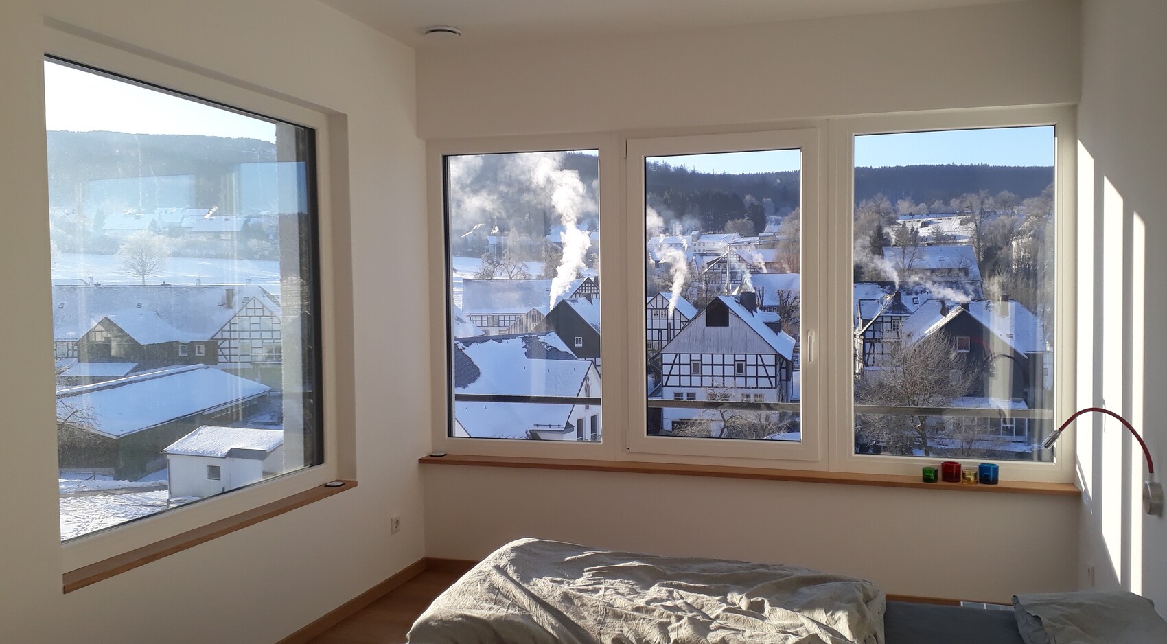 Heller Schlafraum mit großen Fenstern; verschneite Häuser, Rauch aus Schornsteinen, Bett vorn.