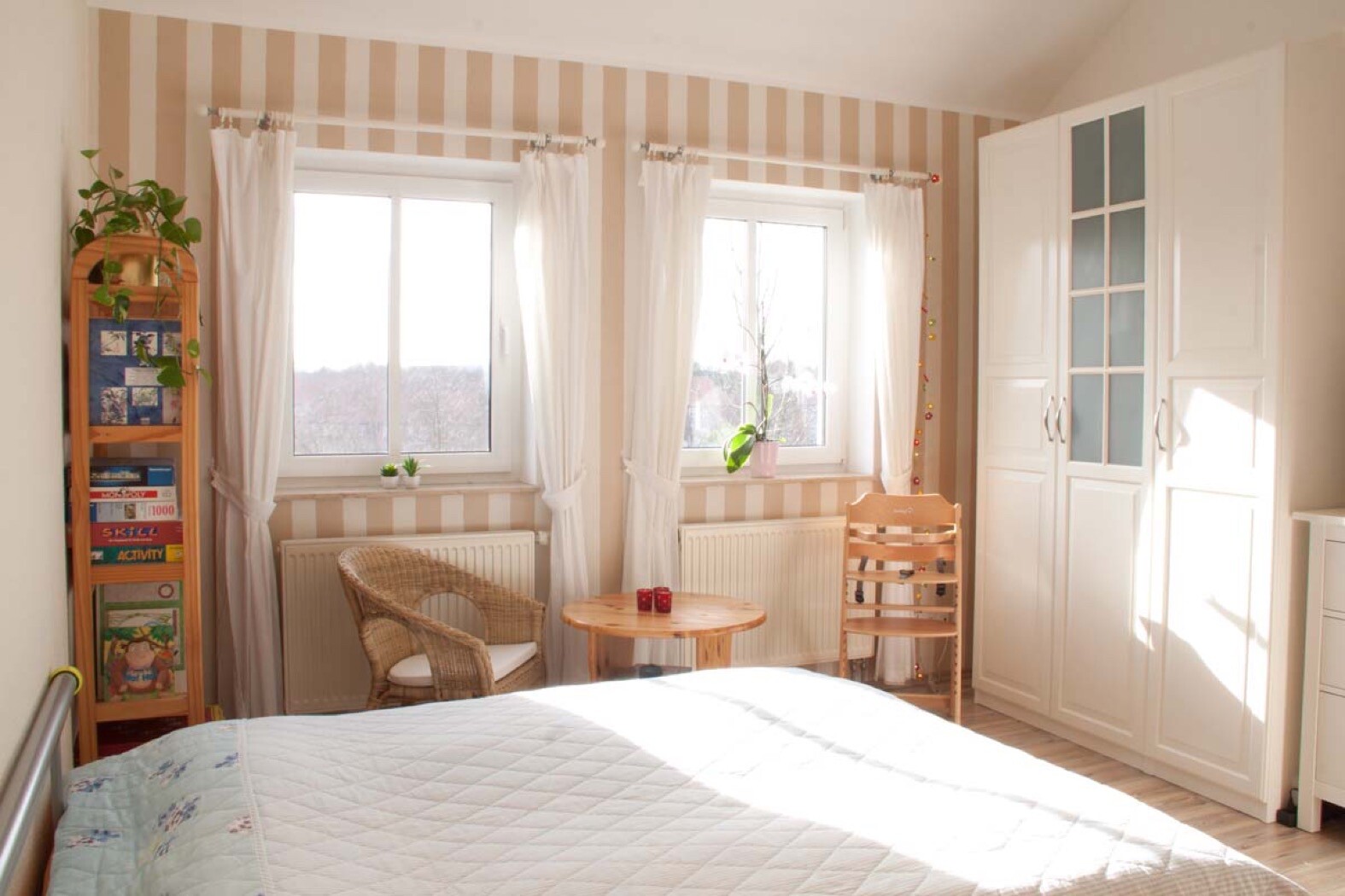 Helles Schlafzimmer mit Bett im Vordergrund, zwei Fenster, runder Holztisch und Kleiderschrank.