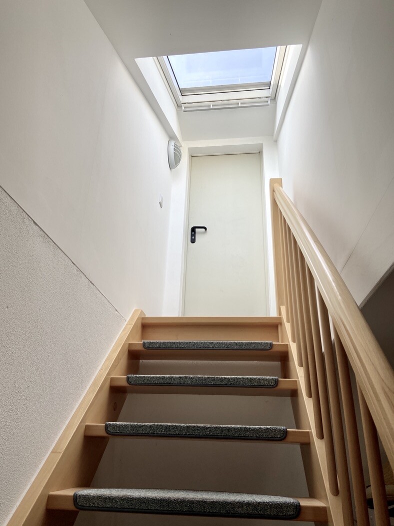 Holztreppe mit rutschfesten Stufen, Geländer rechts, Tür am oberen Ende unter Dachfenster.