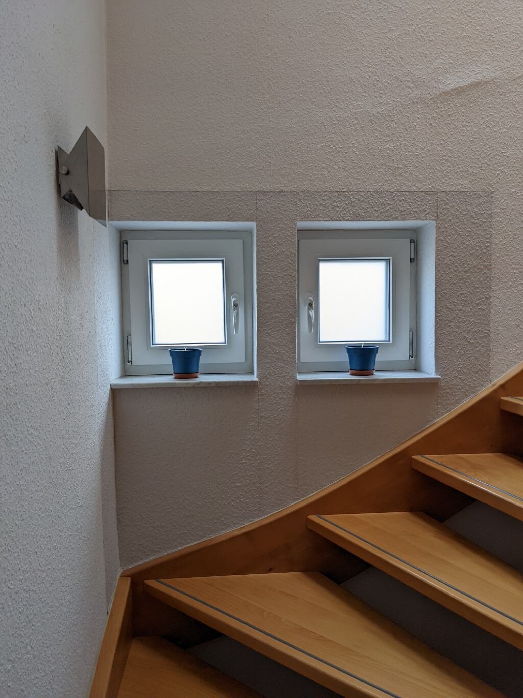 Zwei quadratische Fenster im Treppenhaus mit blauen Blumentöpfen auf der Fensterbank; Holzstufen.
