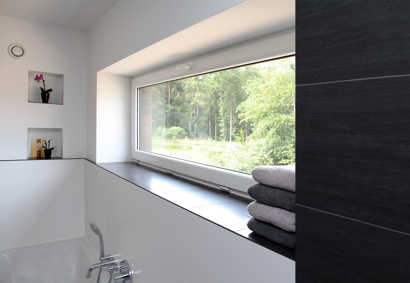 Helles Badezimmer mit Fensterfront, Blick ins Grüne, Badewanne, weiße Fliesen, Handtücher.
