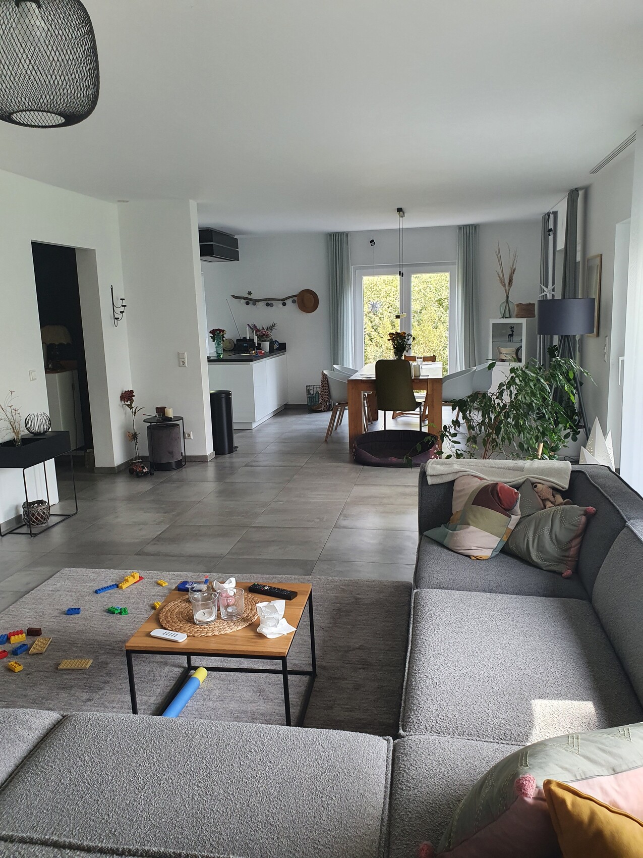 Offenes Wohnzimmer: graue Eckcouch, Lego-Steine auf Tisch, Esstisch mit Stühlen, Pflanzen.