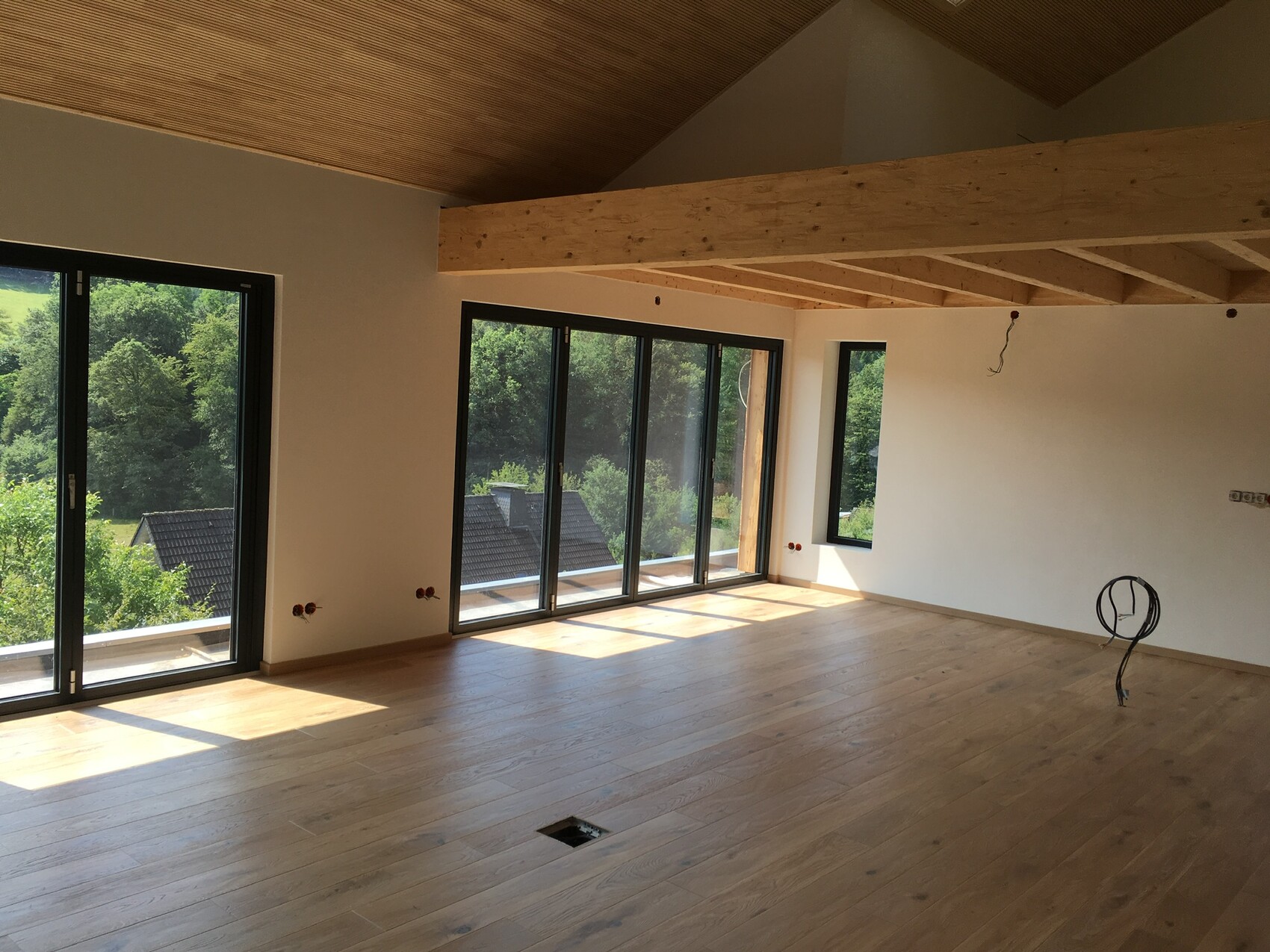 Heller unfertiger Wohnraum mit Holzboden, großen Glasfronten und offener Holzbalkendecke.