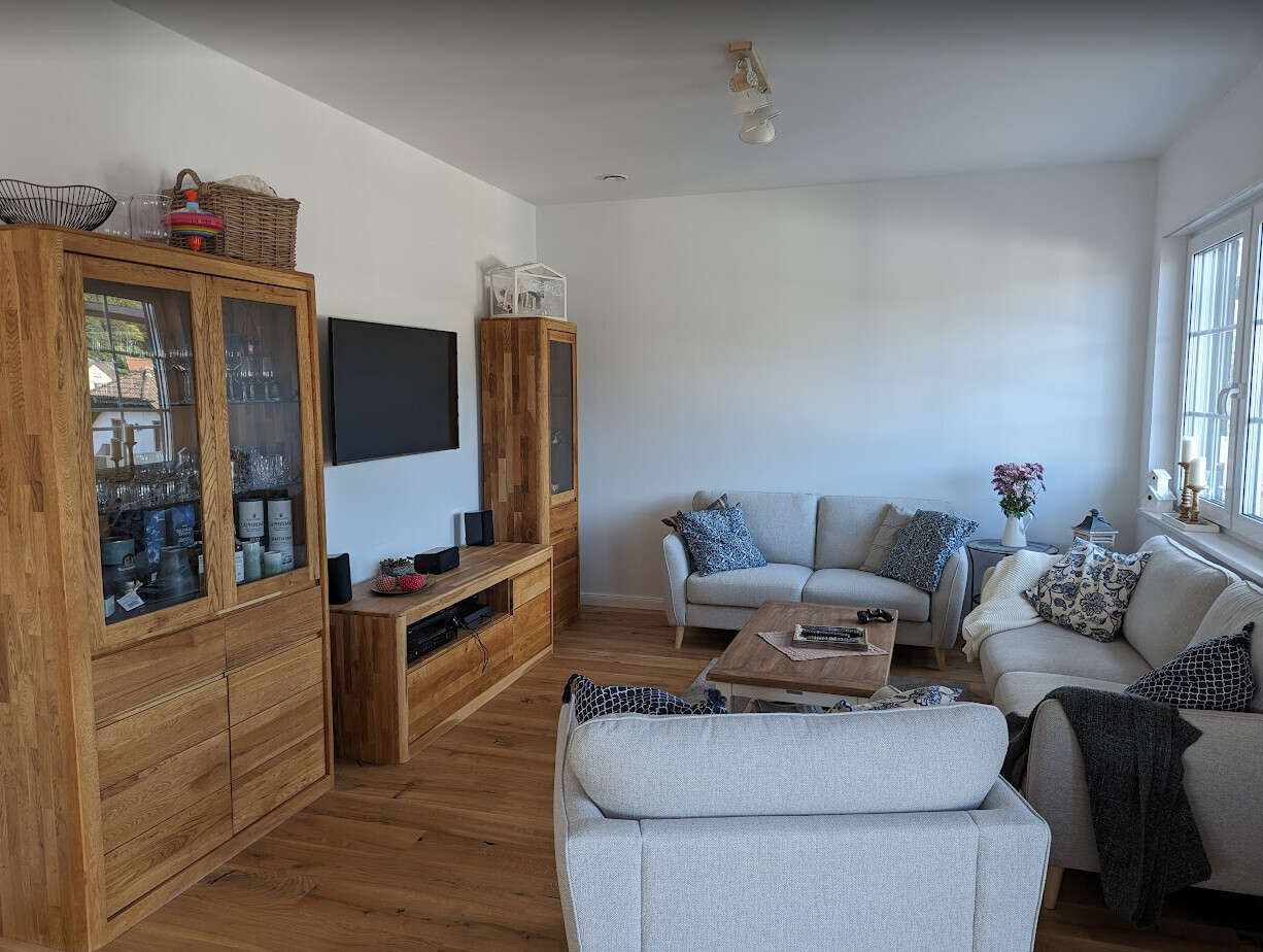 Helles Wohnzimmer mit grauen Sofas, Holzschränken, TV an der Wand, Couchtisch und großen Fenstern.
