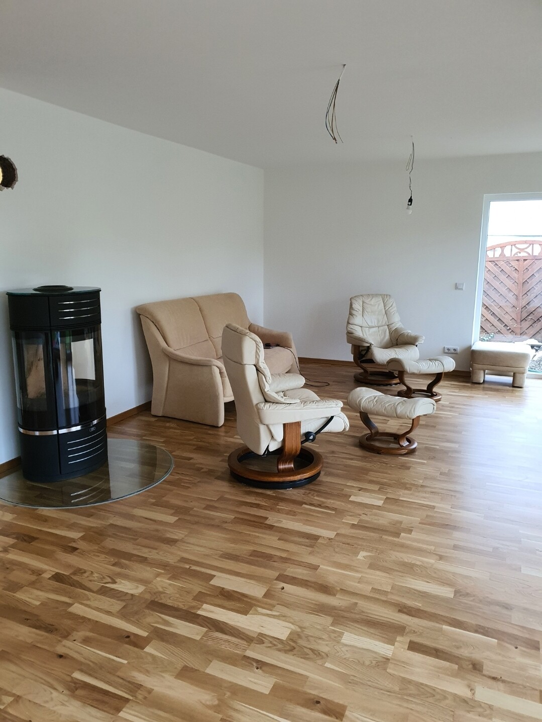 Wohnzimmer mit Holzboden, beige Sessel, Kaminofen; lose Kabel von Decke; Tür zum Garten.