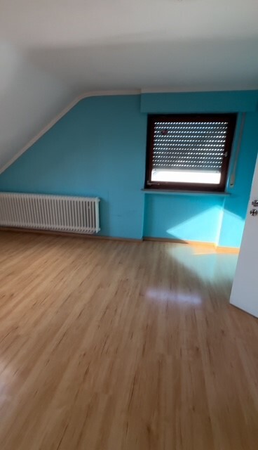 Leeres Zimmer mit Holzboden, blauer Wandfarbe, Heizkörper links, Fenster mit Rollladen rechts.