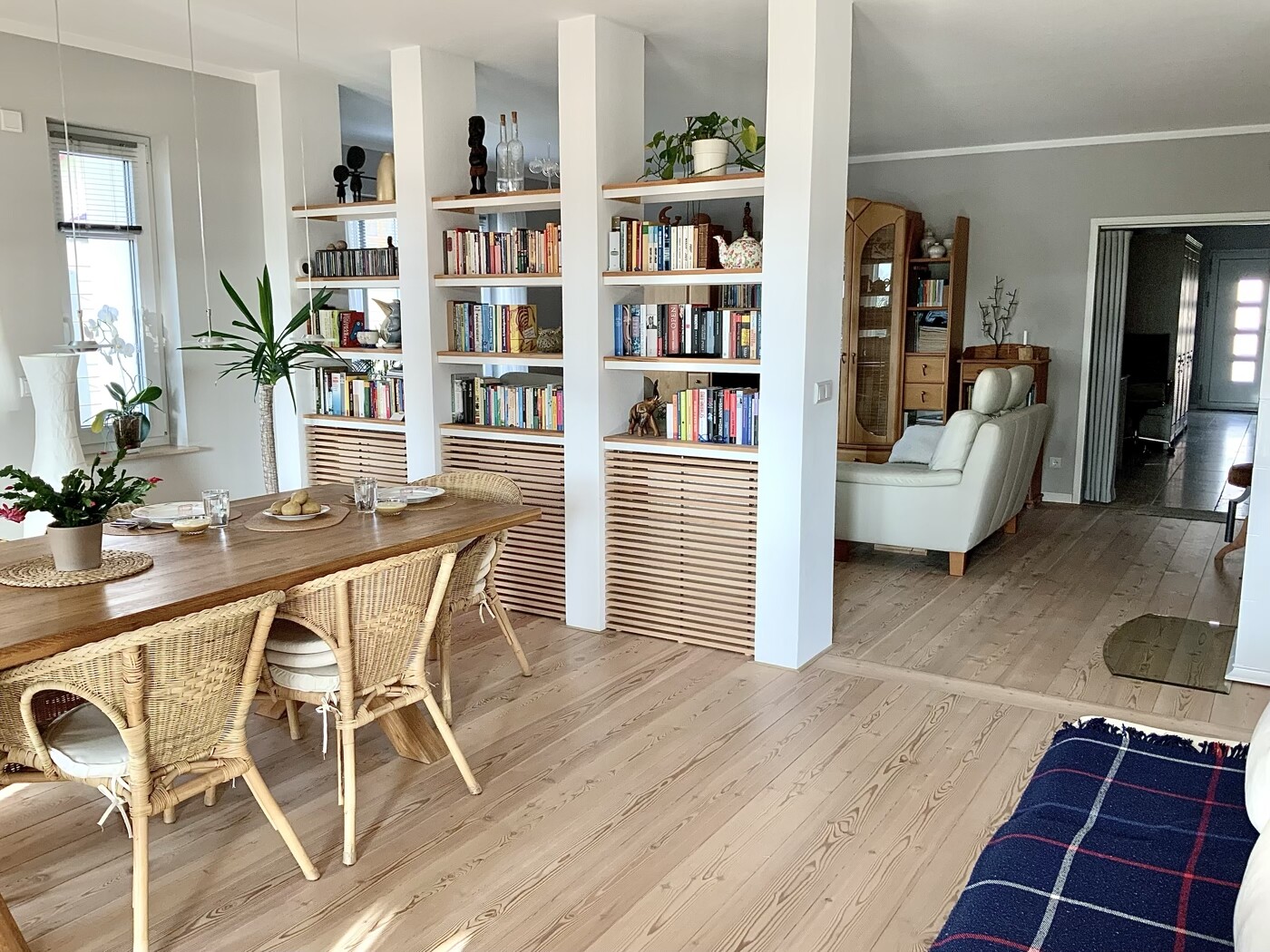 Heller Wohn- und Essbereich mit Holztisch, Korbsesseln, Regal-Raumteiler, Pflanzen und Sofa.