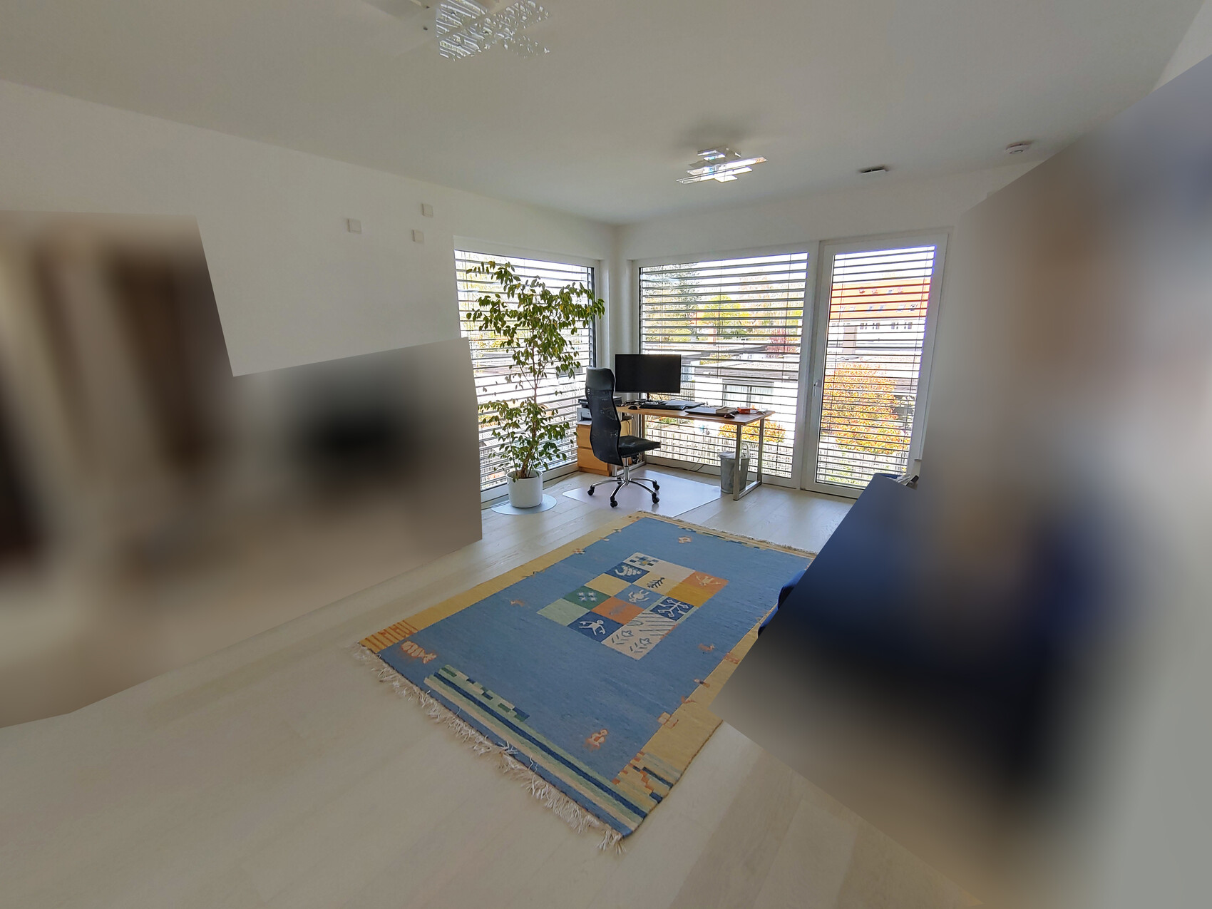 Helles Arbeitszimmer mit großer Fensterfront, Schreibtisch, Monitor, Bürostuhl und Grünpflanze.