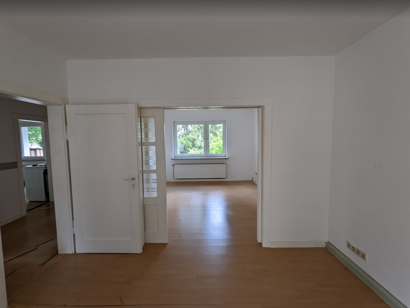 Helle leere Wohnräume mit Holzboden, offener Türrahmen und Fenster mit Gartenblick.