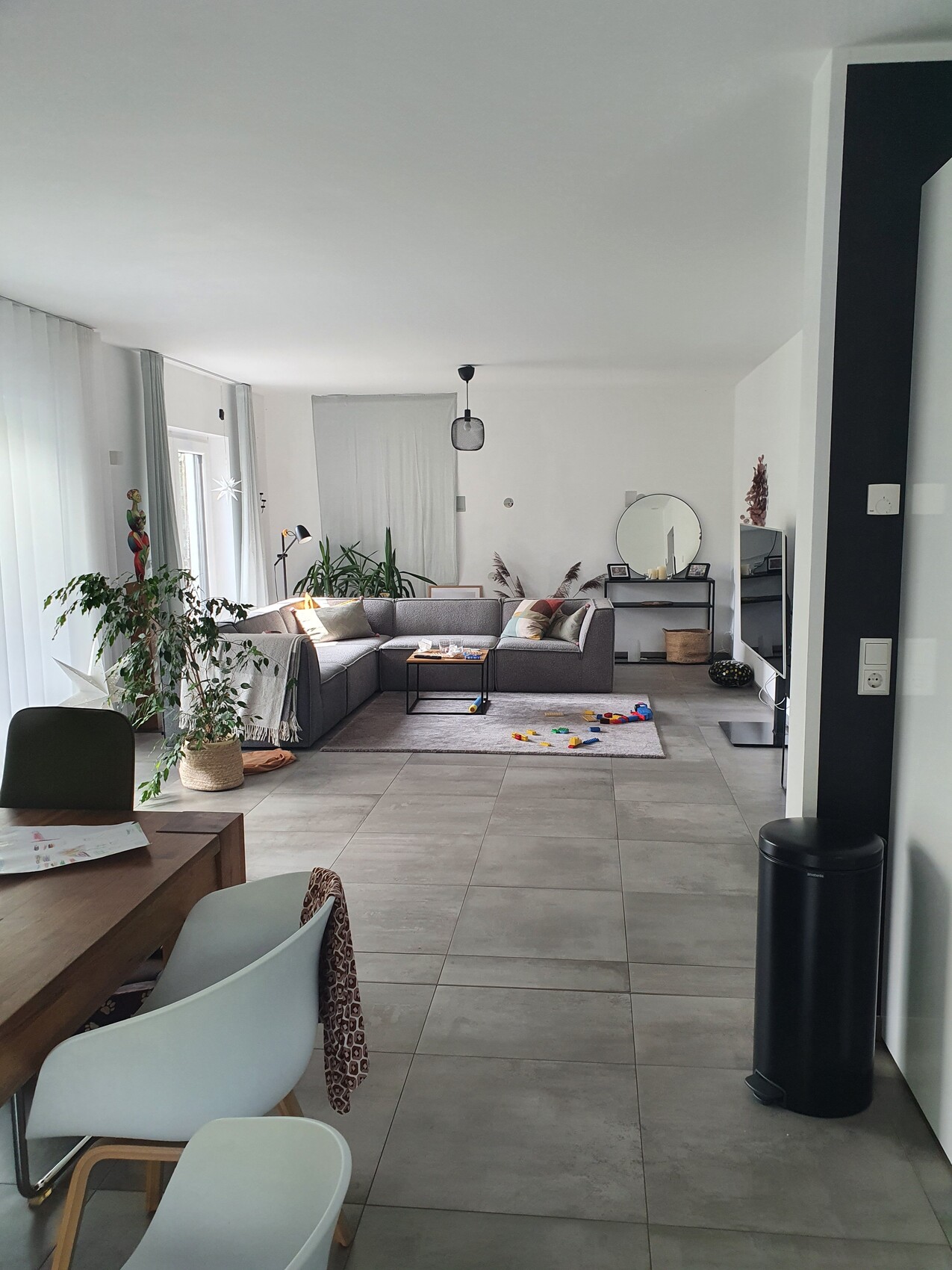 Offenes Wohnzimmer mit grauer Sofaecke, Esstisch links, Pflanzen und Spielzeug auf Boden.