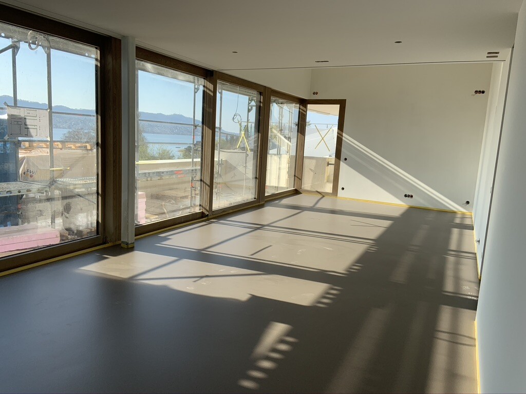 Leerer Innenraum mit großen Fenstern, Blick auf See und Berge, Sonne wirft Schatten.