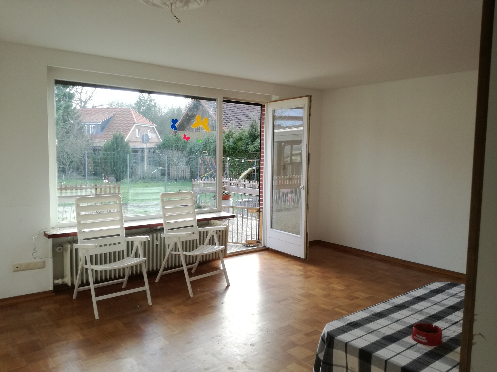 Helles Wohnzimmer mit zwei weißen Klappstühlen am Fenster, offene Tür nach draußen mit Gartenblick.
