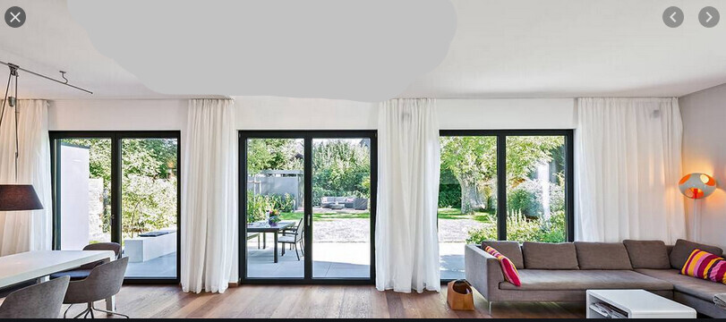 Wohnzimmer mit großen Glas-Schiebetüren, graues Sofa, bunte Kissen, Gartenblick.