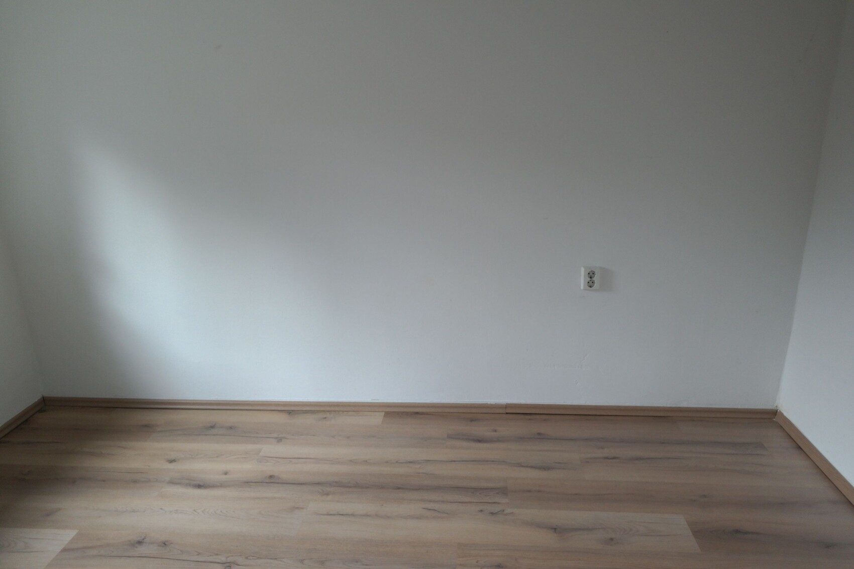 Leerer Innenraum in einer Ecke: weiße Wände, Holzboden, Steckdose an der Wand.