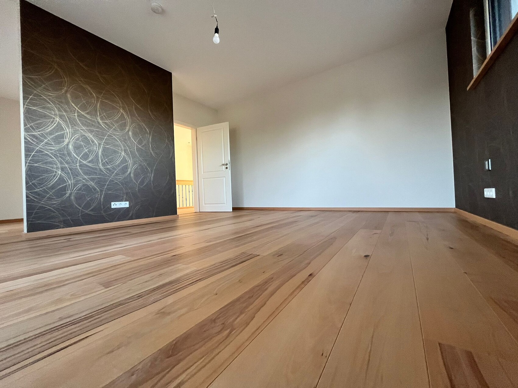 Leeres Zimmer mit Holzboden, linke dunkle Wand mit Spiralmuster, offene Tür zum Flur.