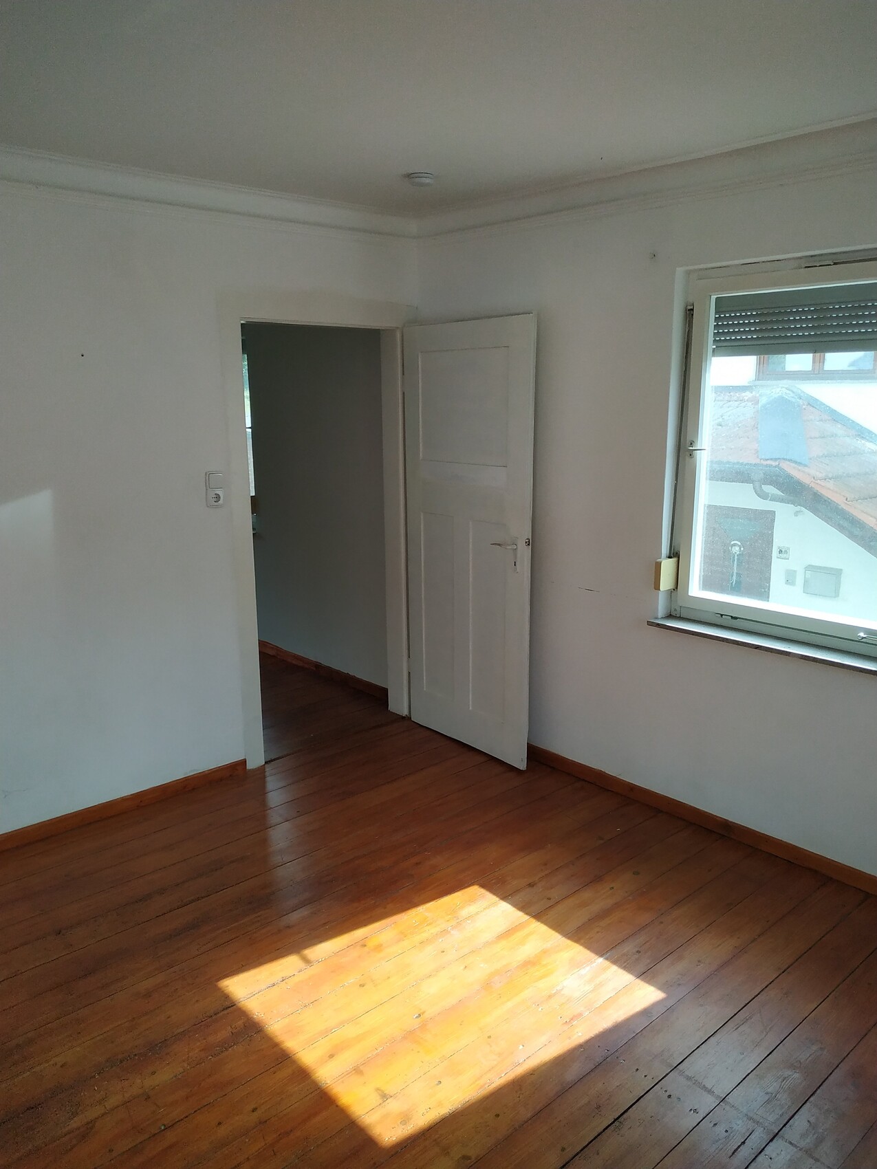 Leerer Raum mit Holzboden, offener Tür, Fenster rechts, Sonnenlichtfleck auf dem Boden.