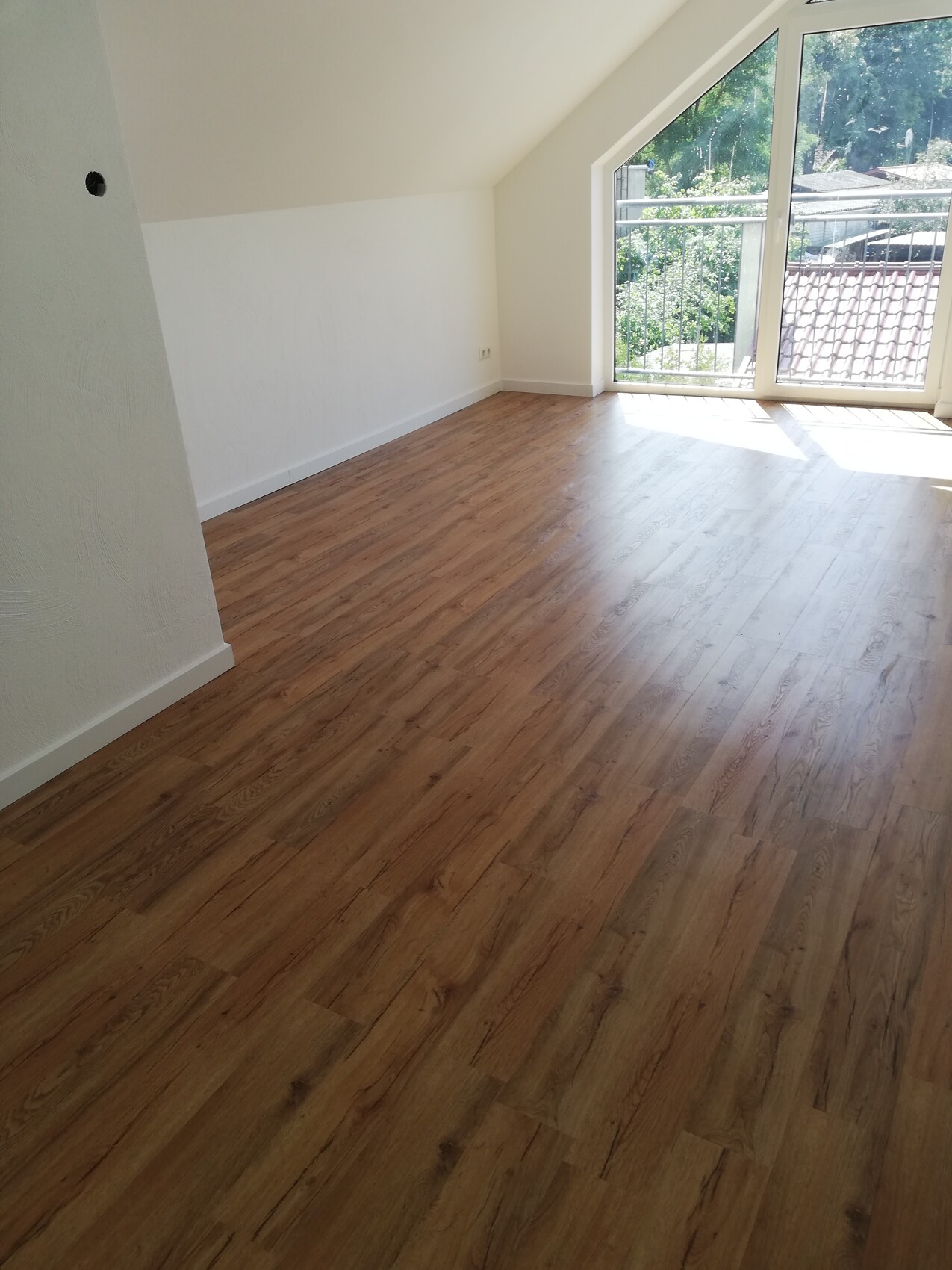 Helles leeres Zimmer mit Holzboden, weißen Wänden und großen Fenstern mit Balkonblick.