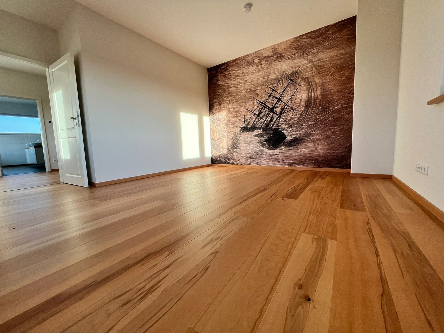 Leerer Wohnraum mit hellem Holzboden; Wandbild eines Segelschiffs an der Rückwand.