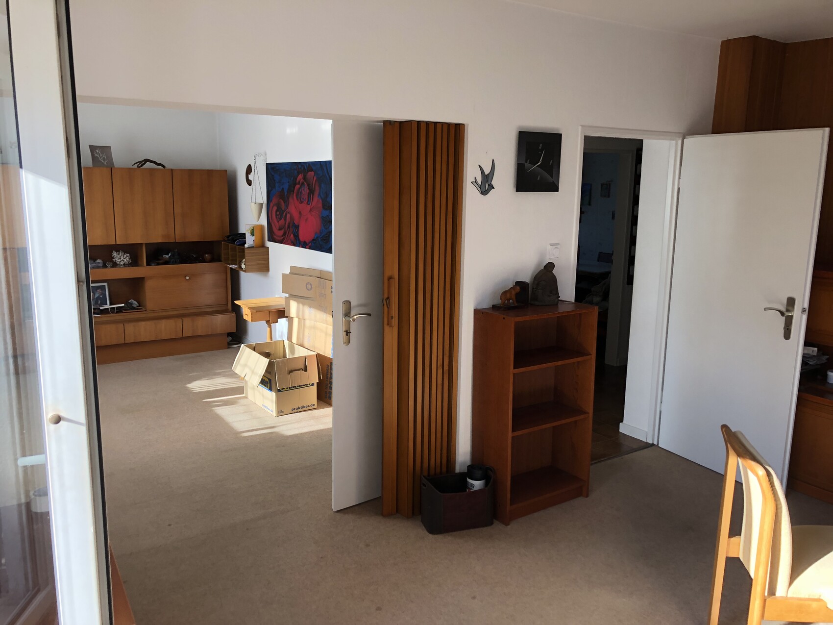 Wohnzimmer mit Holzmöbeln, offener Tür, Kartonbox auf dem Boden, Rosen-Wandbild.