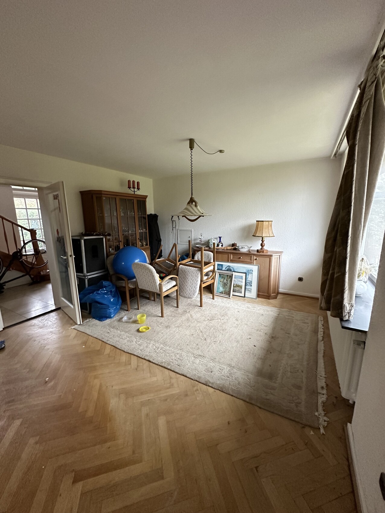 Helles Wohnzimmer mit Sitzgruppe, Vitrinenschrank, Bilderrahmen, Deckenlampe, Fenster mit Vorhängen.