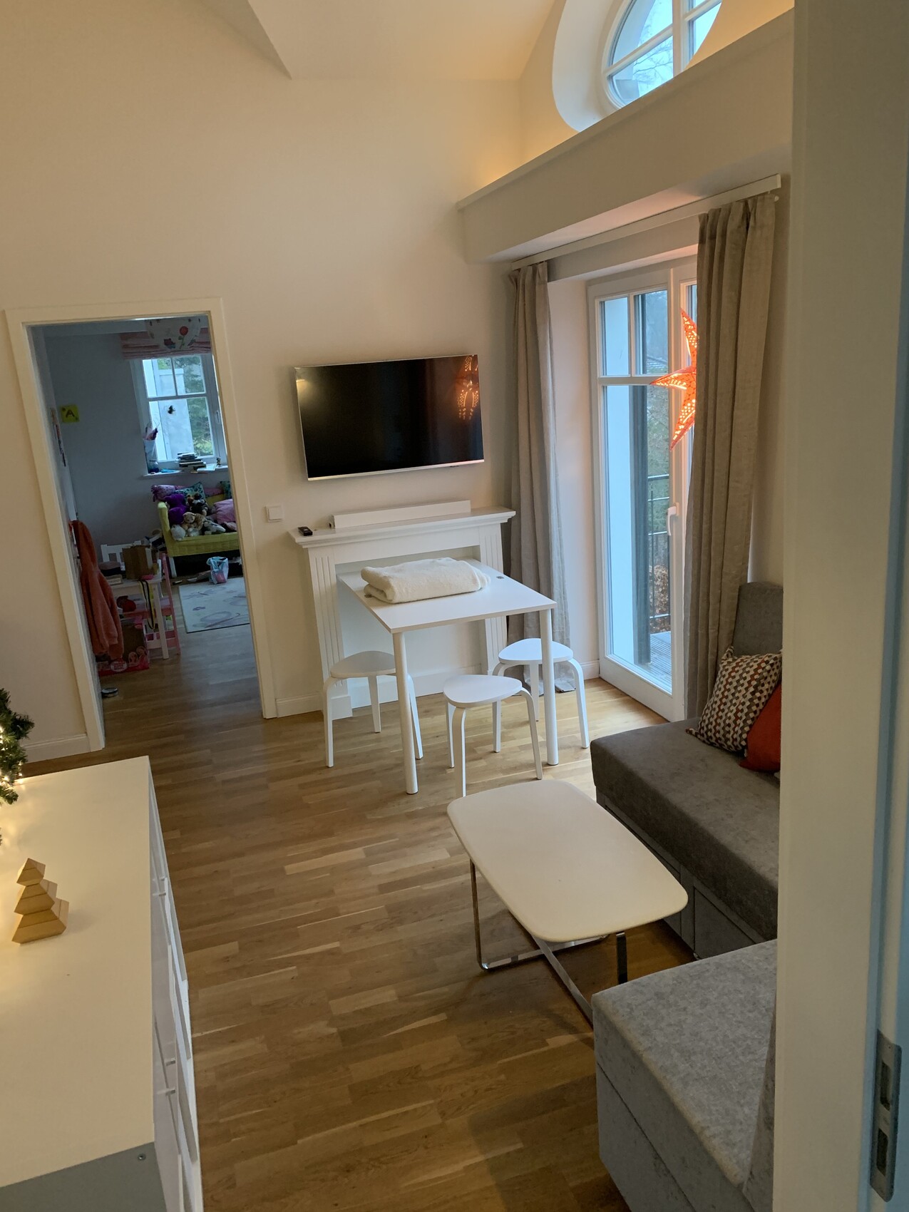 Wohnzimmer mit Fernseher an der Wand, Esstisch mit drei Hockern, graues Sofa, Fenster.