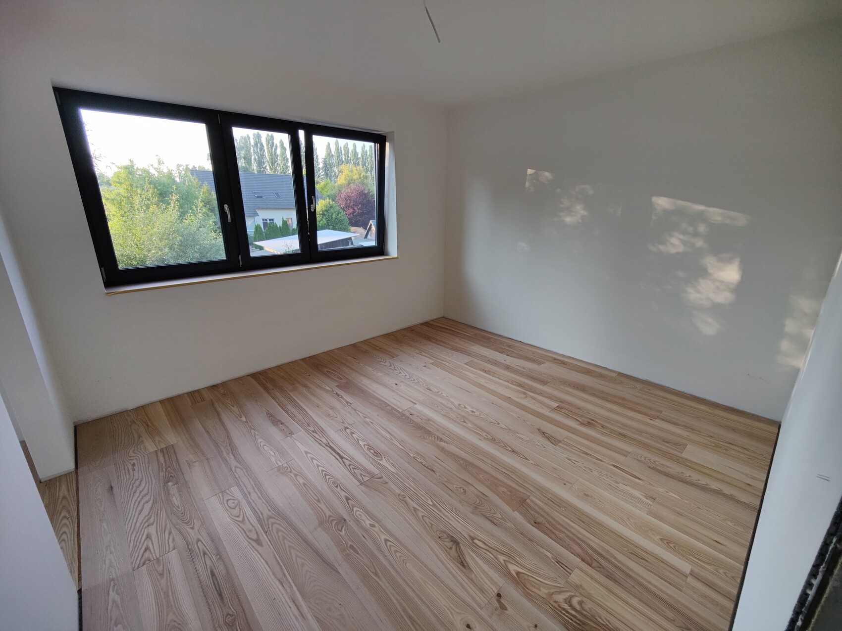 Leeres Zimmer mit hellen Wänden, Holzfußboden und großem dreiteiligem Fenster mit Blick auf Bäume.
