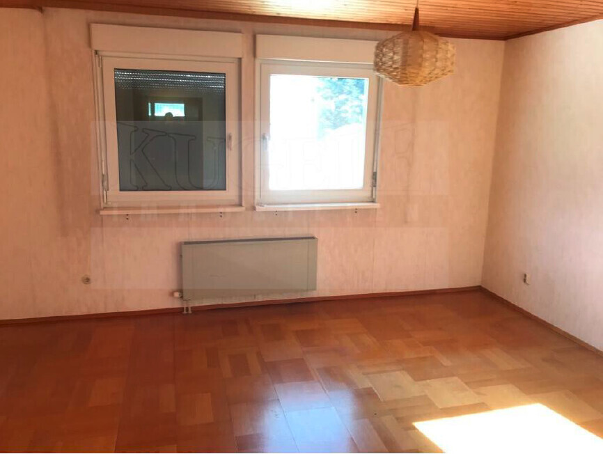 Leeres Zimmer mit Holzparkett, zwei Fenster, Heizkörper darunter und hängender Lampenschirm.