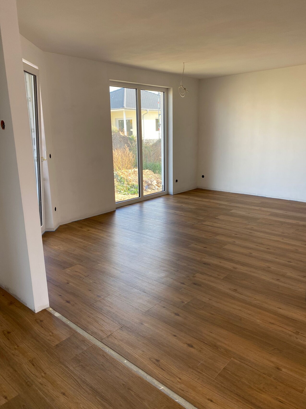 Heller, leerer Wohnraum mit Holzfußboden und großer Glas-Schiebetür nach draußen.