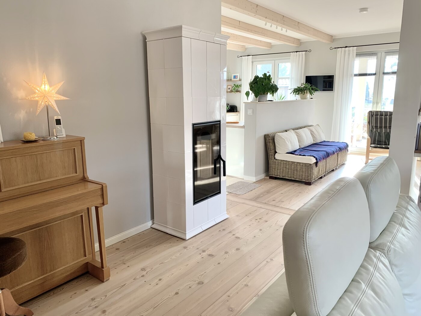 Helles Wohnzimmer mit weißem Kachelofen, Korbsessel-Sofa, Holzfußboden und großen Fenstern.
