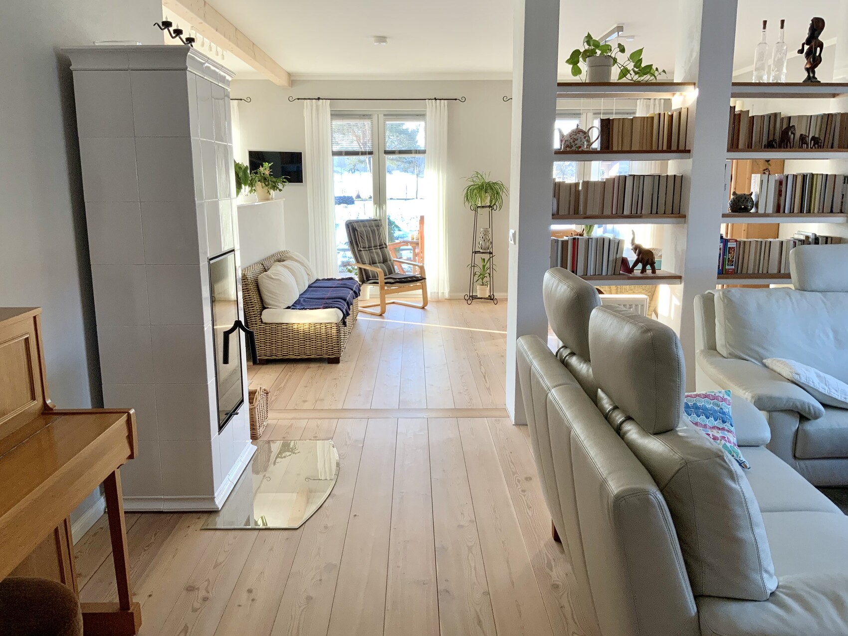 Helles Wohnzimmer mit Kachelofen, Holzboden, Sofa und Sessel sowie einer Bücherregal-Trennwand.