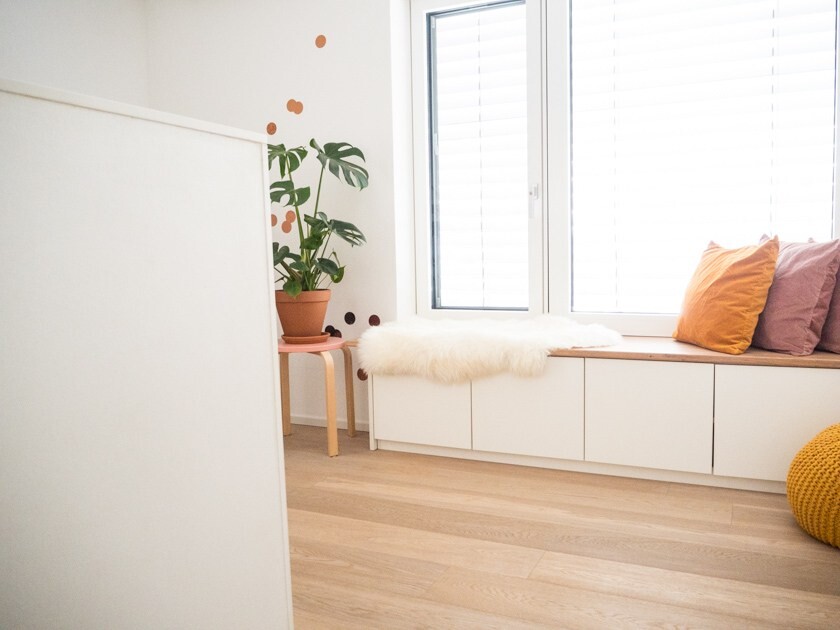 Helles Wohnzimmer mit Fensterbank-Sitzbank, Kissen, Fell-Decke und Pflanze.