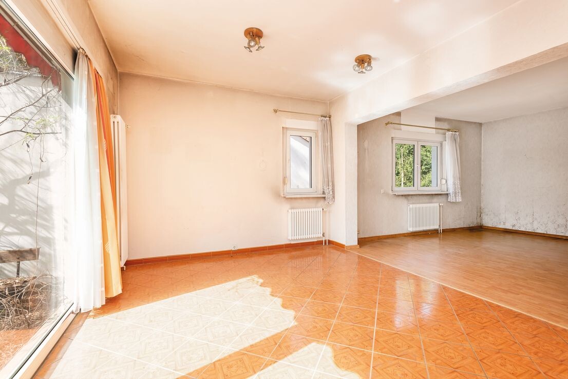 Helles Wohnzimmer mit Terrassenzugang, großen Fenstern, Radiatoren und Fliesenboden.